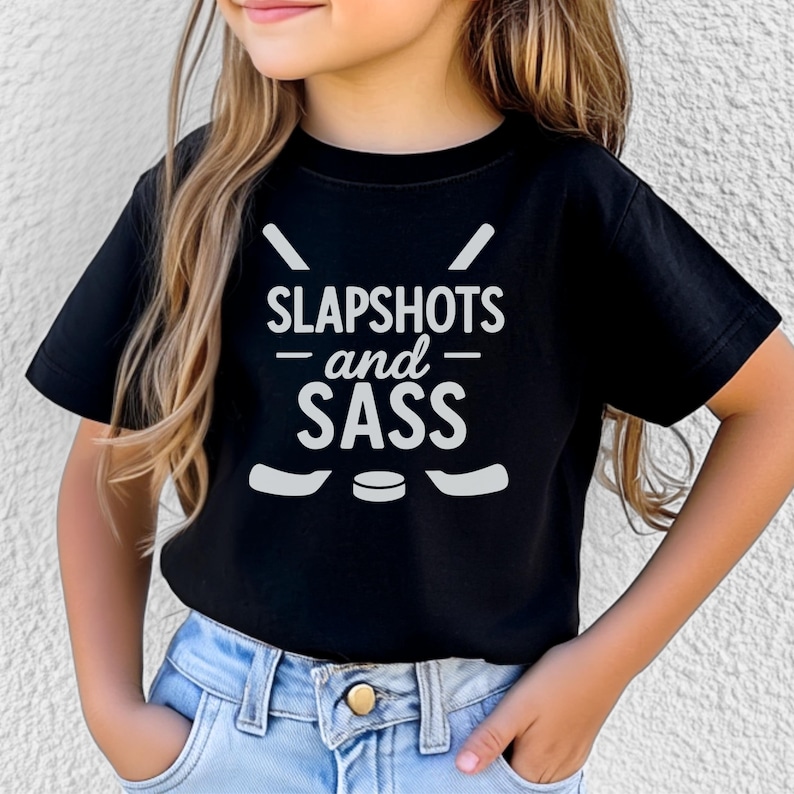 Op de afbeelding: Zwart T-shirt met de witte tekst "SLAPSHOTS and SASS". Het ontwerp bevat hockeysticks en een puck. Het shirt wordt door een kind gedragen.
