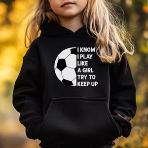May include: A black hooded sweatshirt featuring a white graphic of a soccer ball on the left side and the text "I KNOW I PLAY LIKE A GIRL TRY TO KEEP UP" in white capital letters on the right side, including a front kangaroo pocket.