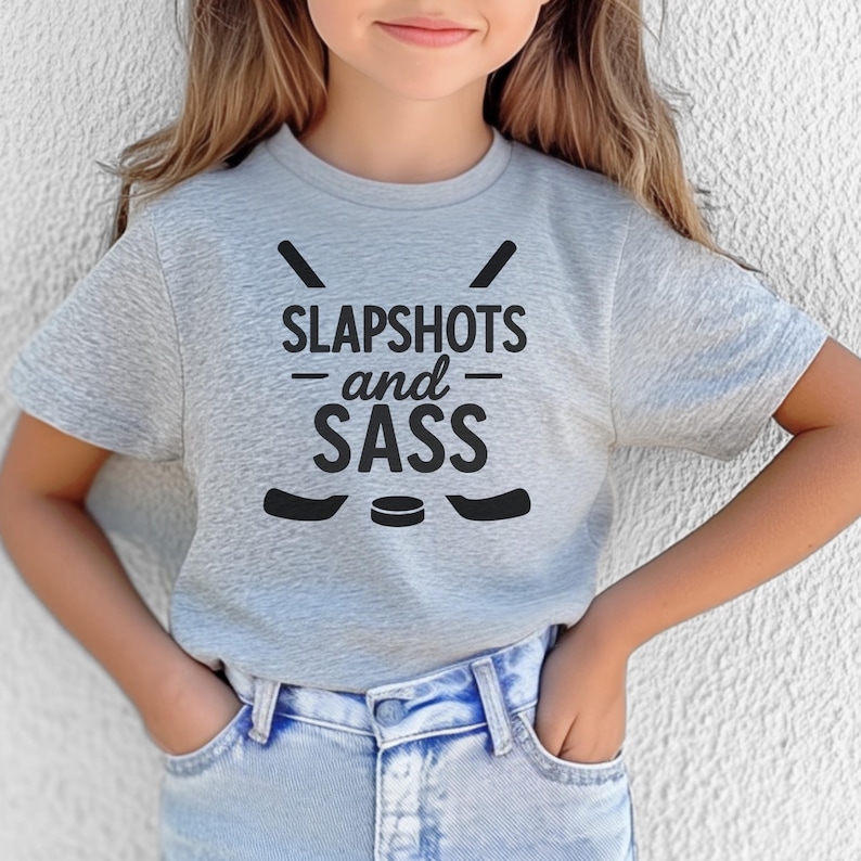 Op de afbeelding: Een lichtgrijs t-shirt met de zwarte tekst "SLAPSHOTS and SASS", samen met hockeysticks en een puck-afbeelding. Het shirt wordt gedragen door een persoon die een blauwe spijkerbroek draagt.