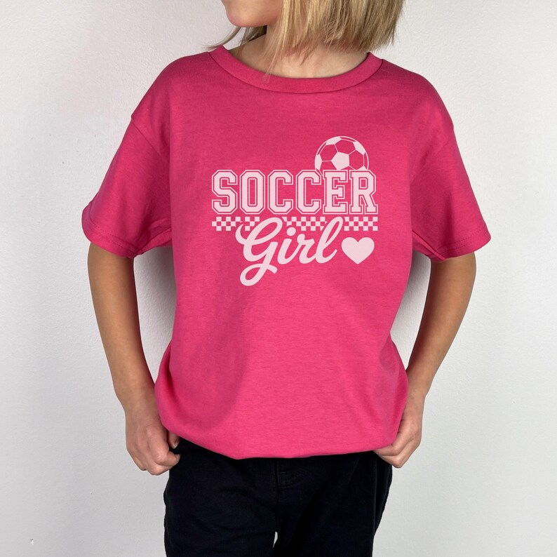May include: A magenta t-shirt with the words "SOCCER Girl" in white, featuring a soccer ball graphic and a heart. The shirt is being worn by a person, with black pants.