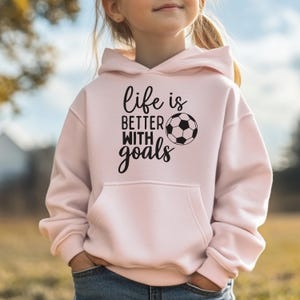 May include: A light pink hooded sweatshirt featuring a front kangaroo pocket and black text that reads "life is BETTER WITH goals" alongside a graphic illustration of a soccer ball. The design is centered on the chest of the long-sleeved garment.