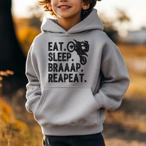 May include: A light gray hoodie with a front pocket and the words "EAT. SLEEP. BRAAAP. REAPEAT." in black, along with a graphic of a motorcycle. The hoodie is worn by a person.