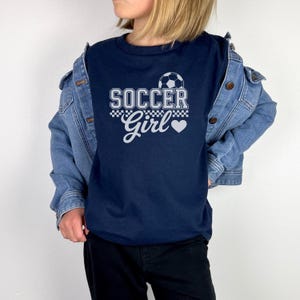May include: Navy blue t-shirt with the text "SOCCER Girl" in white, featuring a soccer ball graphic and a heart. The shirt is worn with a denim jacket and black pants.