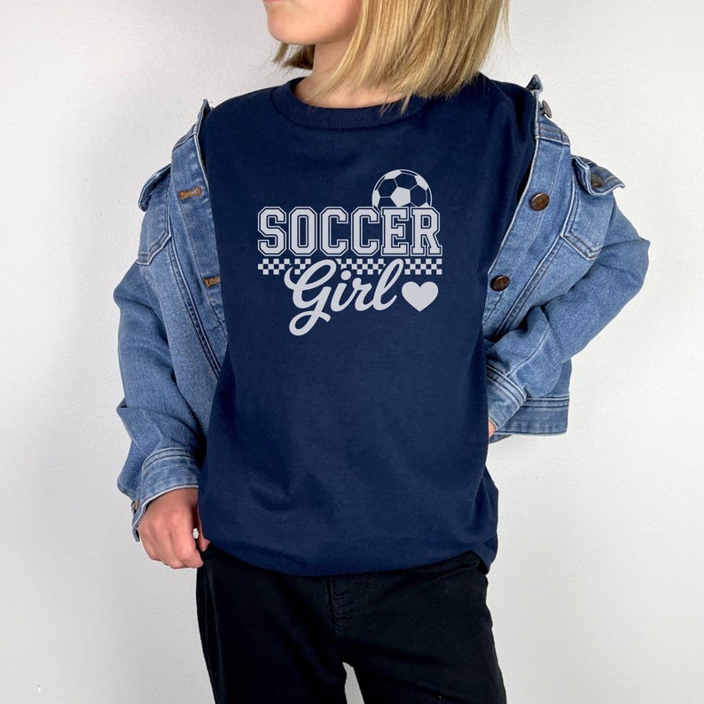 May include: Navy blue t-shirt with the text "SOCCER Girl" in white, featuring a soccer ball graphic and a heart. The shirt is worn with a denim jacket and black pants.