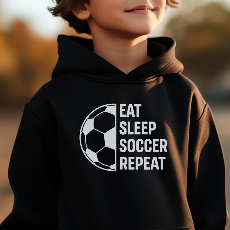 May include: Black hooded sweatshirt with a white soccer ball graphic and the words "EAT SLEEP SOCCER REPEAT". The sweatshirt is made of a soft material and has a drawstring hood.