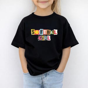 May include: A black t-shirt with the words "SOFTBALL GIRL" in a colourful, collage-style design. The shirt features a softball graphic and is worn by a person wearing blue jeans.