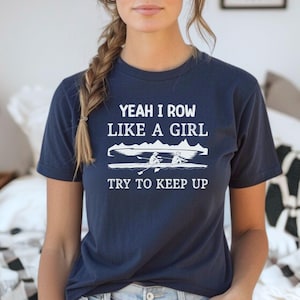 May include: A navy blue t-shirt with white text that reads "Yeah I row like a girl try to keep up". The shirt features a graphic of two people rowing a boat.