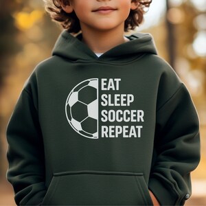 May include: Dark green hoodie with a white soccer ball graphic and the text "EAT SLEEP SOCCER REPEAT". The hoodie has a front pocket and a hood. The image is of a child wearing the hoodie.