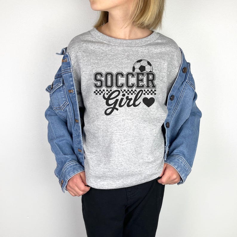 May include: A gray long-sleeve shirt with the words "SOCCER Girl" in black and white. A soccer ball graphic is above the word "SOCCER". The word "Girl" is in a script font with a black heart.