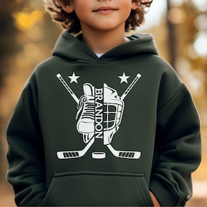 May include: Dark green hoodie featuring a white hockey design. The graphic includes a hockey skate, helmet, crossed hockey sticks, a puck, and the name "BRANDON". Two white stars complete the design.