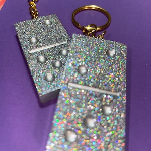 May include: Two glittery domino keychains. Each rectangular charm is silver with iridescent glitter and white dots. The keychains have gold-colored key rings and chains. The background is a solid purple.