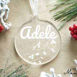 May include: White acrylic Christmas ornament with the name "Adele" written in a cursive font and a white floral design.