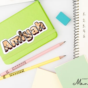 Peut inclure: Un carnet vert avec un autocollant qui dit "Amiyah" en lettres à carreaux brunes et blanches. Le carnet est sur une surface blanche avec deux crayons et une gomme bleue.