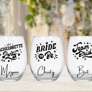 May include: Three clear glass wine glasses with black lettering. The glasses say "Bachelorette Party" with the name Megan, "Bride to Be" with the name Christy, and "Team Bride" with the name Brit.