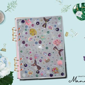 May include: A clear plastic binder with a mermaid theme. The binder has a variety of colorful embellishments, including seashells, starfish, and mermaids. The binder is closed and has gold rings.