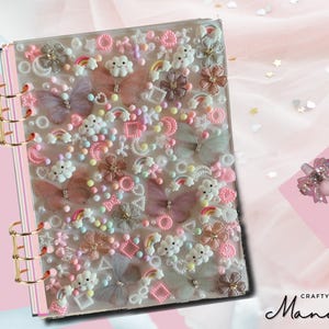 Puede incluir: Un archivador rosa y blanco con una cubierta transparente decorada con varios adornos, incluyendo nubes, arcoíris, mariposas y perlas. El archivador tiene un sistema de encuadernación de anillos dorados.