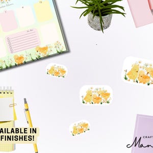 May include: A flat lay featuring a weekly planner, stickers, and stationery. The stickers depict yellow chicks with floral accents. A yellow pen and notepad are also visible. Text reads "AVAILABLE IN 5 FINISHES!"