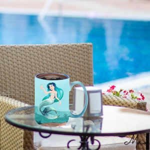 Könnte beinhalten: Eine blaue Keramiktasse mit einem Meerjungfrauen-Motiv. Die Tasse ist mit einer braunen Flüssigkeit gefüllt und hat einen weißen Henkel. Die Tasse steht auf einem Glastisch mit einem Korbstuhl im Hintergrund.