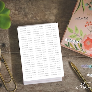 Op de afbeelding: Een verzameling stickers en briefpapier op een houten oppervlak. De stickers tonen de tekst "CUSTOM TEXT" in een herhalend patroon. Een bloemrijk notitieboek en een gouden pen zijn ook zichtbaar. Het woord "customtext" staat op het notitieboek.