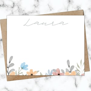 May include: A white card with the name "Laura" written in cursive at the top. The bottom of the card is decorated with a border of watercolour flowers in shades of blue, pink, and orange.