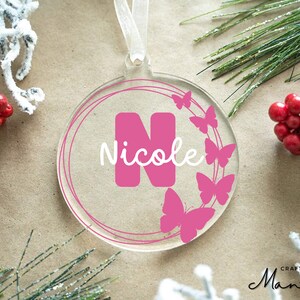May include: Pink acrylic Christmas ornament with a white ribbon, featuring a large pink letter N and the name "Nicole" in white script, surrounded by a wreath of pink butterflies.