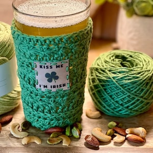 Pu&ograve; includere: Un copri-birra in crochet verde con un'etichetta che dice "Kiss Me I'm Irish" e un trifoglio. Il copri-birra &egrave; su un bicchiere di birra con una testa di schiuma. Ci sono filati verdi e noci su un tavolo di legno.