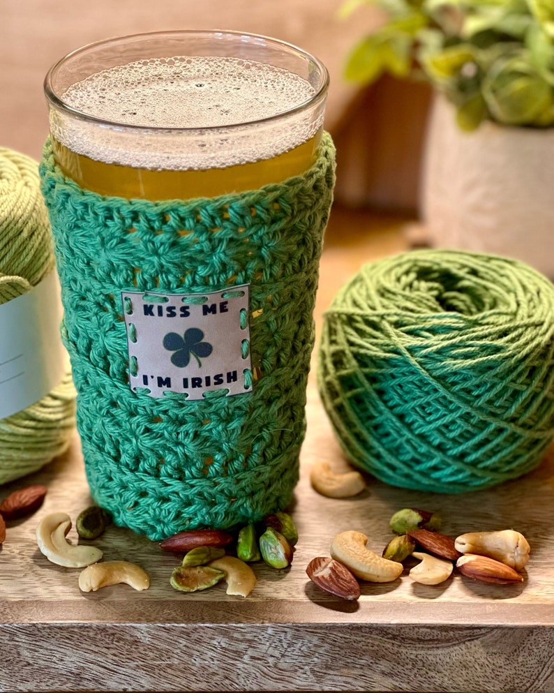 Pu&ograve; includere: Un copri-birra in crochet verde con un'etichetta che dice "Kiss Me I'm Irish" e un trifoglio. Il copri-birra &egrave; su un bicchiere di birra con una testa di schiuma. Ci sono filati verdi e noci su un tavolo di legno.