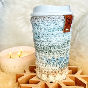 Pu&ograve; includere: Un copri-tazza da caff&egrave; all'uncinetto in tonalit&agrave; di blu, verde e beige con un'etichetta in pelle marrone. Il copri-tazza &egrave; appoggiato su un sottobicchiere di legno a forma di fiocco di neve.