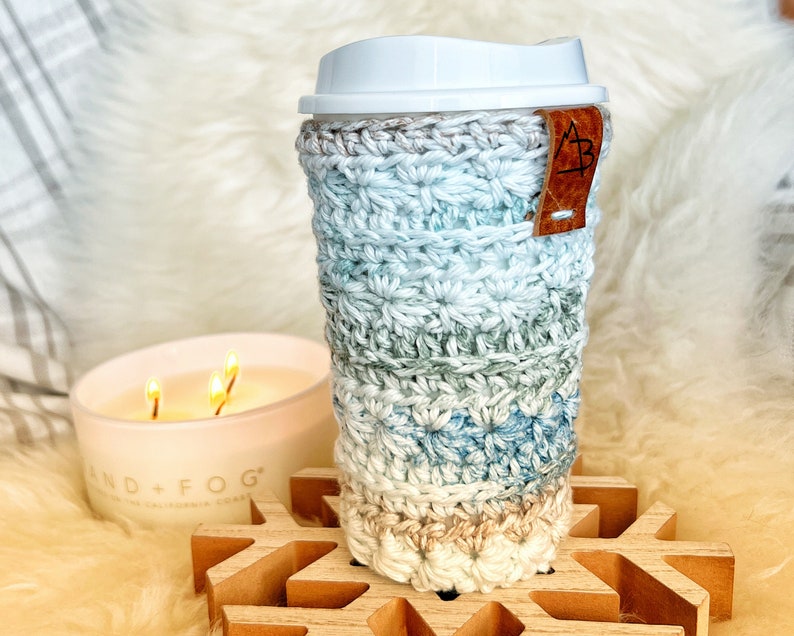 Pu&ograve; includere: Un copri-tazza da caff&egrave; all'uncinetto in tonalit&agrave; di blu, verde e beige con un'etichetta in pelle marrone. Il copri-tazza &egrave; appoggiato su un sottobicchiere di legno a forma di fiocco di neve.