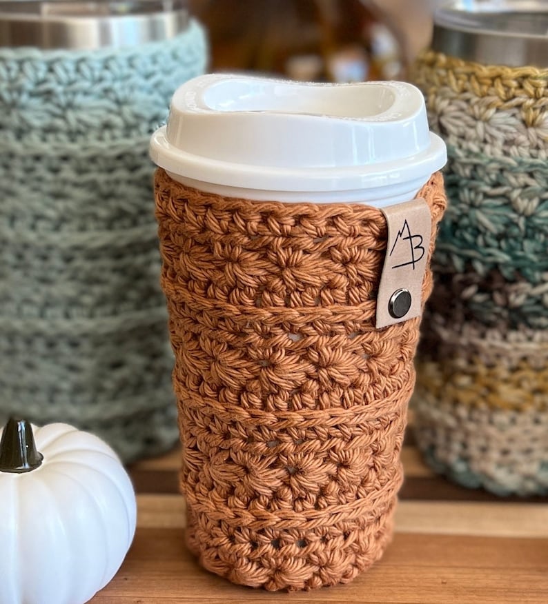 Pu&ograve; includere: Un copri-tazza da caff&egrave; all'uncinetto di colore arancio bruciato con un cinturino in pelle marrone con le iniziali "A B".