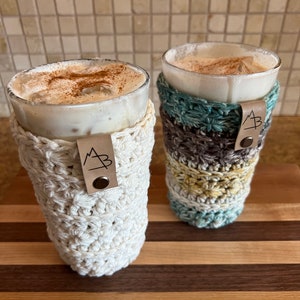Pu&ograve; includere: Due bevande al caff&egrave; freddo in bicchieri trasparenti con fodere all'uncinetto. Le fodere sono bianche e multicolori con un'etichetta in pelle marrone che dice "MB" su ciascuna. Le bevande sono su un tagliere di legno.