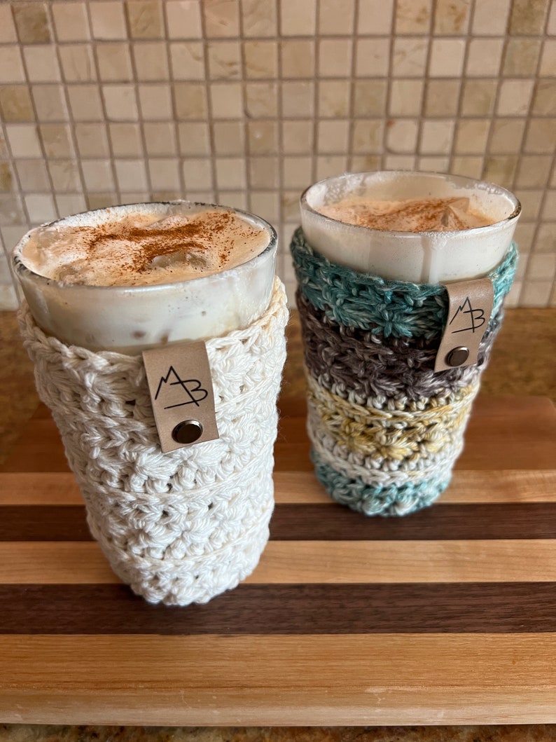 Pu&ograve; includere: Due bevande al caff&egrave; freddo in bicchieri trasparenti con fodere all'uncinetto. Le fodere sono bianche e multicolori con un'etichetta in pelle marrone che dice "MB" su ciascuna. Le bevande sono su un tagliere di legno.