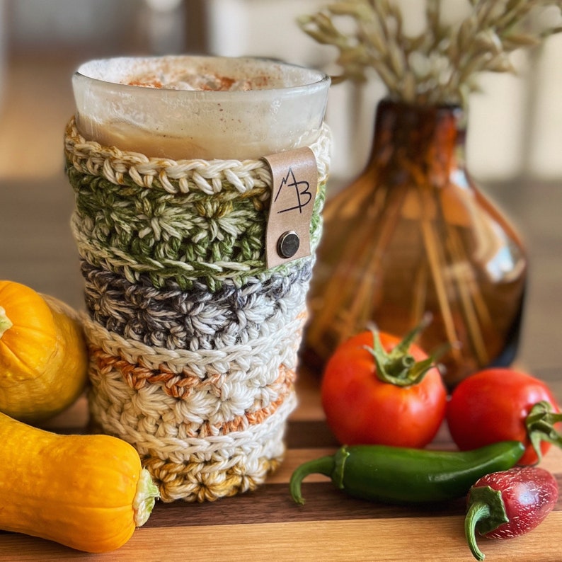 Pu&ograve; includere: Un copri-tazza da caff&egrave; all'uncinetto con un cinturino in pelle marrone e la lettera "A" su di esso. Il copri-tazza &egrave; verde, grigio e beige. La tazza &egrave; piena di una bevanda marrone chiaro.