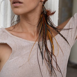 Puede incluir: Una mujer con un turbante marrón y una blusa beige con largos pendientes de plumas. Los pendientes son marrones, negros y dorados.