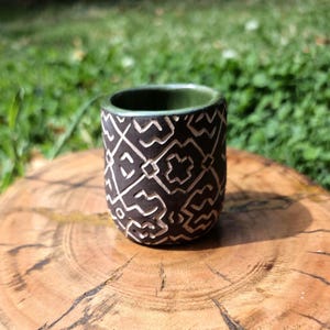 May include: A small, cylindrical ceramic cup featuring a dark brown exterior with a carved, light-coloured geometric pattern of interlocking lines and shapes. The interior of the cup is finished in a smooth, solid olive green glaze.