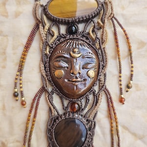 May include: A handcrafted macrame necklace featuring a central face pendant with closed eyes, adorned with gold accents. The necklace incorporates brown and tan cords, along with oval and round tiger's eye stones, creating a bohemian aesthetic.