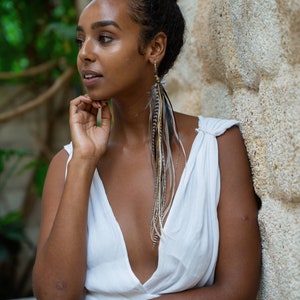May include: A woman with dark brown hair in a bun wears a white sleeveless dress with a deep V-neckline. She is wearing long feather earrings with brown, black, and white feathers.