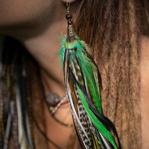 May include: A pair of long, green and black feather earrings with a gold bead and a small, green and gold charm. The earrings are hanging from a person's ear.