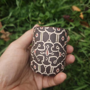 May include: A cylindrical dark brown ceramic cup featuring a repeating geometric pattern carved into the surface. The intricate design is highlighted with thin white lines and small red rectangular accents, creating a textured, symmetrical motif.