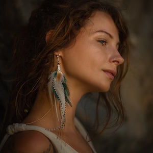 Könnte beinhalten: Eine Frau mit langen braunen Haaren trägt ein weißes ärmelloses Oberteil und einen langen Federohrring. Der Ohrring besteht aus weißen, braunen und blauen Federn.