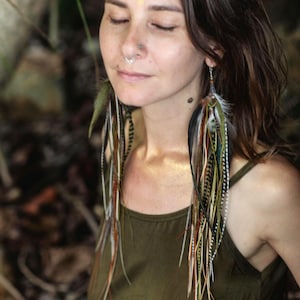 May include: A woman with long brown hair wearing a green tank top and long feather earrings. The earrings are multi-colored with brown, white, and gold feathers.