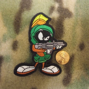May include: Embroidered patch featuring the cartoon character Marvin the Martian, holding a weapon. The patch is green, orange, and black with white accents. A gold coin is next to the patch. The background is camouflage.