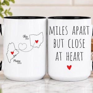 May include: Two white ceramic mugs with black handles and rims. One mug features black outlines of the states of Maine and Ohio with red hearts and a dashed line connecting them, labeled with names. The other mug displays the text MILES APART BUT CLOSE AT HEART with a red heart.