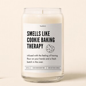 May include: A clear glass jar candle filled with white wax. The label reads: Tumblerfy, SMELLS LIKE COOKIE BAKING THERAPY, infused with the feeling of having flour on your hands and a fresh batch in the oven, 13.75 oz, 60-80 hours burn time, 100% soy wax candle.
