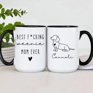 May include: Two white ceramic mugs with black handles and rims. One mug reads "BEST F*CKING weenie MOM EVER" with a heart. The other mug features a dachshund illustration and the name "Cannoli".