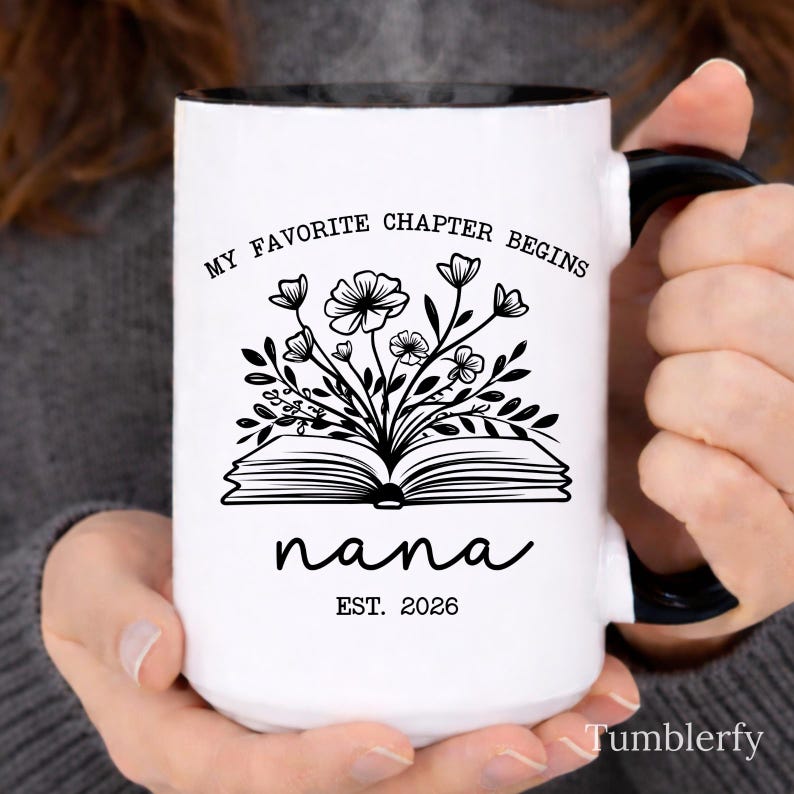 K&ouml;nnte beinhalten: Wei&szlig;er Keramikbecher mit schwarzem Innenraum und Griff. Der Becher tr&auml;gt den Text "MY FAVORITE CHAPTER BEGINS" und "nana EST. 2026" mit einer floralen Illustration von Blumen und Bl&auml;ttern. Der Becher wird von einer Person gehalten.