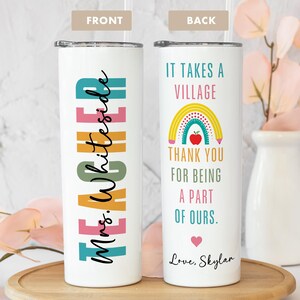 May include: A white tumbler with a lid and straw. The front features the word "TEACHER" in colorful block letters and "Mrs. Whiteside" in cursive. The back says "IT TAKES A VILLAGE" with a rainbow and apple graphic, and "THANK YOU FOR BEING A PART OF OURS."
