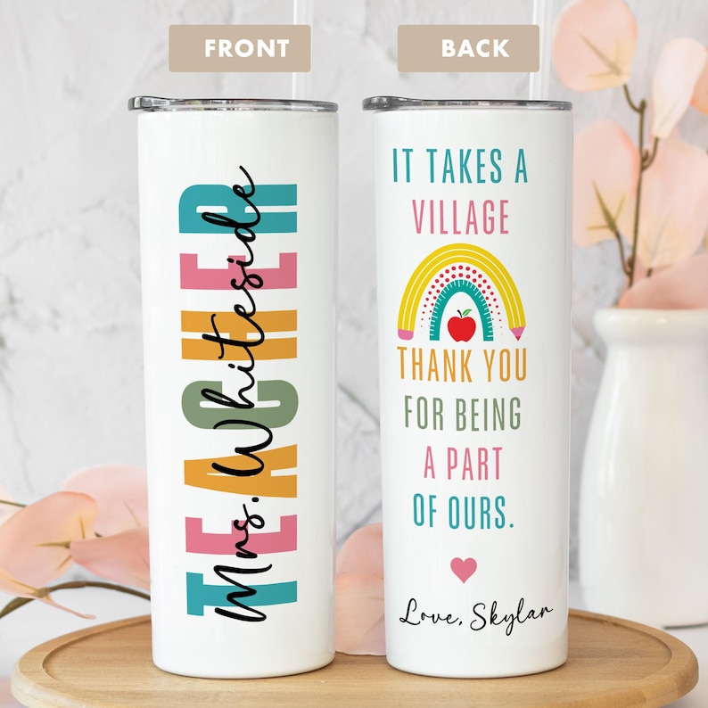 May include: A white tumbler with a lid and straw. The front features the word "TEACHER" in colorful block letters and "Mrs. Whiteside" in cursive. The back says "IT TAKES A VILLAGE" with a rainbow and apple graphic, and "THANK YOU FOR BEING A PART OF OURS."