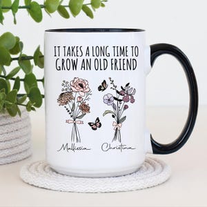 May include: White ceramic mug with a black handle and rim. The mug features the text "IT TAKES A LONG TIME TO GROW AN OLD FRIEND" and two floral bouquets with butterflies. The names "Mallissia" and "Christina" are below the bouquets.