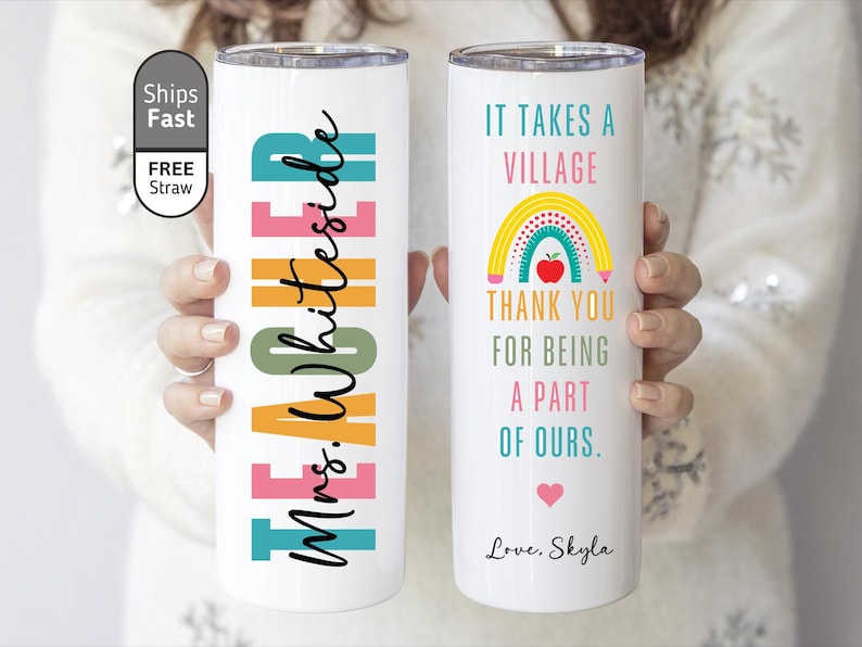 May include: White insulated tumbler with colorful text and graphics. One side reads "Mrs. Whiteside" in black script over colorful blocks. The other side says "It takes a village" with a rainbow and apple graphic. A heart and the name "Skyla" are also included.